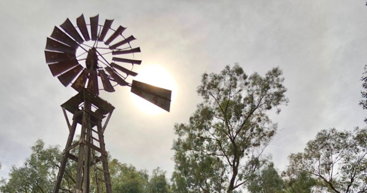 A metal windmill stands tall among trees in a garden, enduring the sun shining through a cloudy sky behind it&mdash;ready to survive summers heat.
