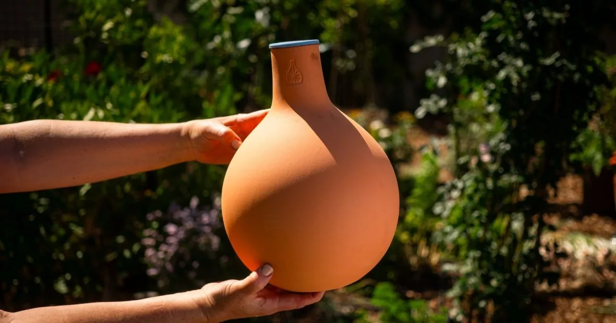 Hands holding a round, orange clay vessel in a sunlit garden, surrounded by green plants—a perfect scene for any gardener's gift guide.