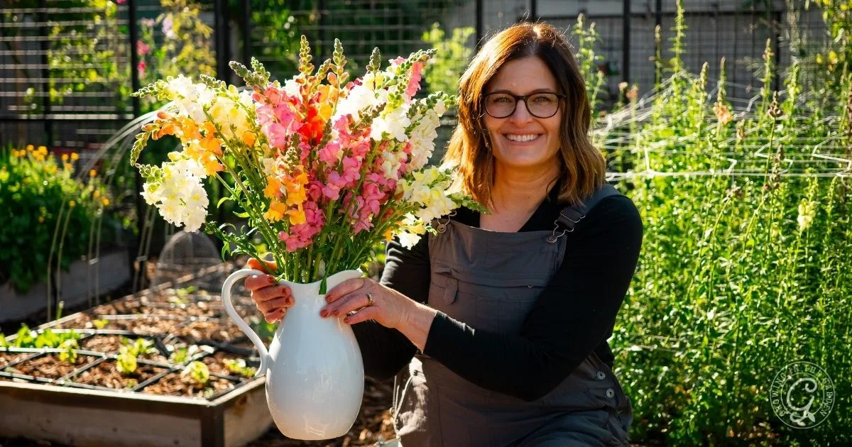 Smiling woman holding a white vase of colorful flowers in a sunny garden, showcasing quick and easy garden flower arrangements.