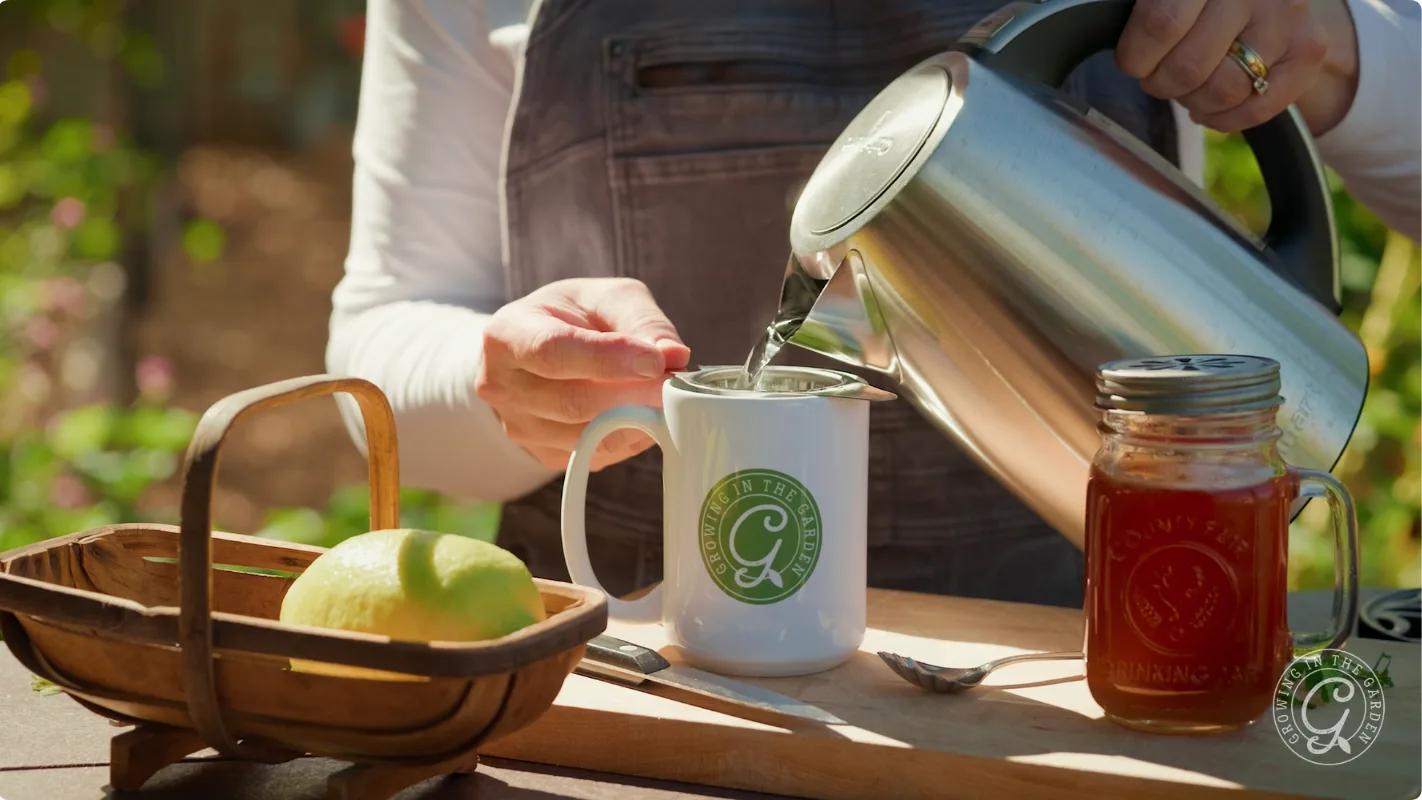 A person pours hot water from a kettle into a mug with a Growing in the Garden logo.