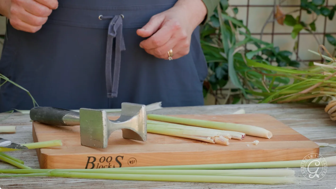 Person standing by a cutting board with lemongrass stalks and a metal meat tenderizer on it.