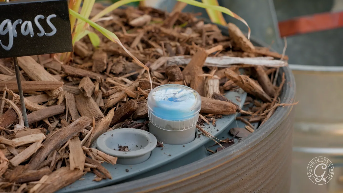 A small blue and white sensor sits in mulched soil next to a gray dish in a garden planter.