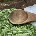 A wooden spoon rests on chopped herbs and coarse salt—perfect essentials for learning how to make & use celery salt—on a rustic wooden surface.