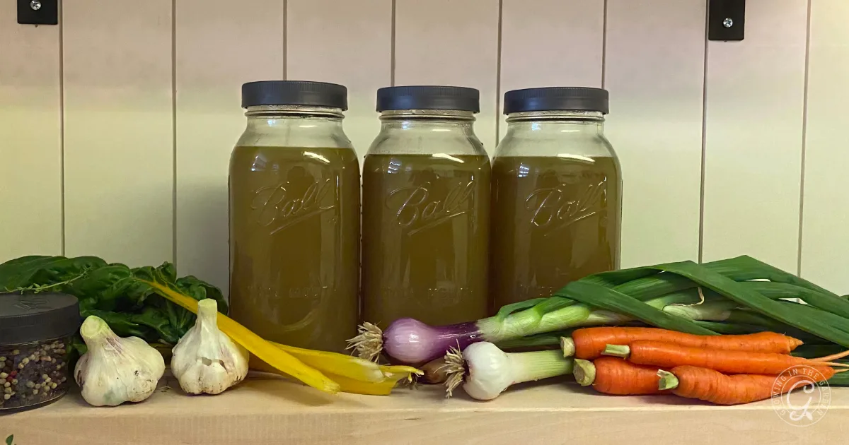 Three jars of garden vegetable broth & stock are displayed on a shelf, surrounded by fresh garlic, carrots, onions, and leafy greens.
