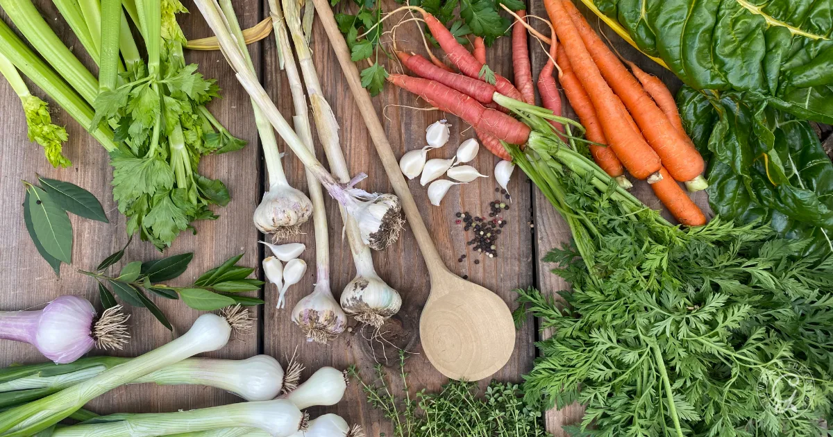Fresh vegetables, herbs, and garlic with a wooden spoon on a rustic wooden table&mdash;perfect ingredients for a garden vegetable broth & stock, seen from above.
