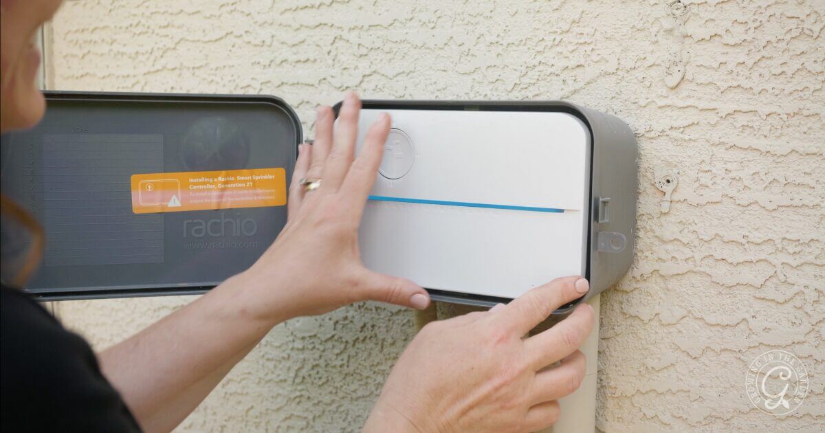 A person installs or adjusts a white outdoor smart irrigation controller mounted on a textured beige wall, with the cover open and hands touching the device.