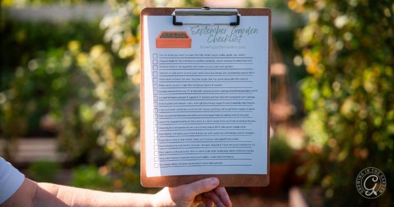 A hand holds a clipboard with a September Garden Checklist in an outdoor garden setting.