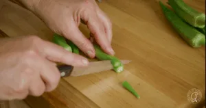 A person slicing the stem off fresh okra pods on a wooden cutting board with a knife, prepping ingredients for the best recipes for okra.