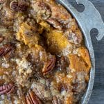 A close-up of a baked pecan and peach dessert in a round metal dish on a wooden surface.