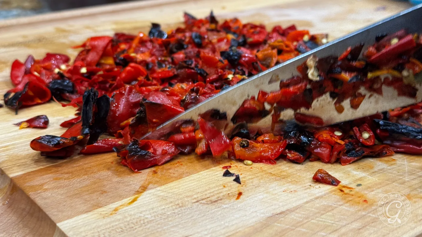 A knife chopping roasted red peppers on a wooden cutting board.