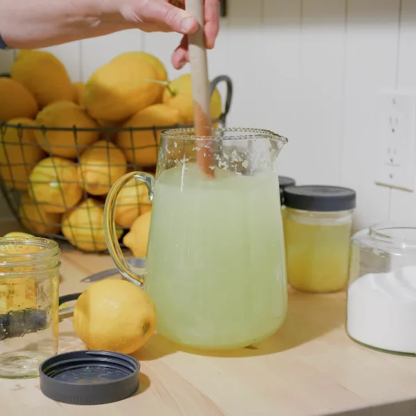 A hand stirs lemonade in a glass pitcher on a counter with lemons, jars, and sugar nearby.