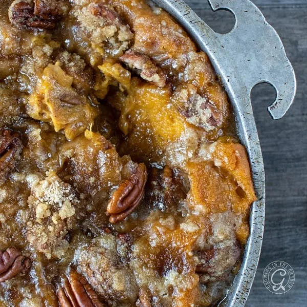 A close-up of pecan-studded sweet potato casserole in a round metal baking dish on a wooden table, next to a platter of Blue Cheese Thyme Scalloped Potatoes.
