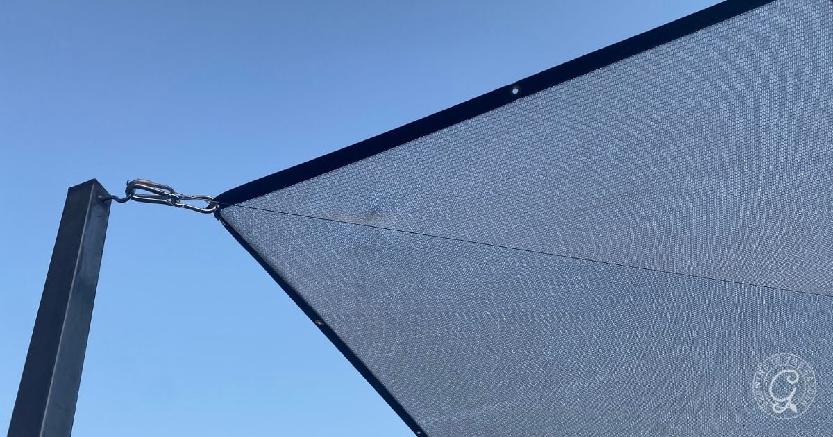 A close-up view of a black triangular sunshade canopy corner, illustrating why adding shade is beneficial. Stretched and attached to a metal pole against a clear blue sky, one corner is secured with a metal ring and clip, showing the texture of the shade fabric.