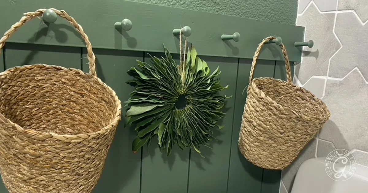 Two wicker baskets and a leafy wreath&mdash;perfect for those curious about how to make a bay laurel wreath&mdash;hang on green wall pegs above a patterned tile floor.