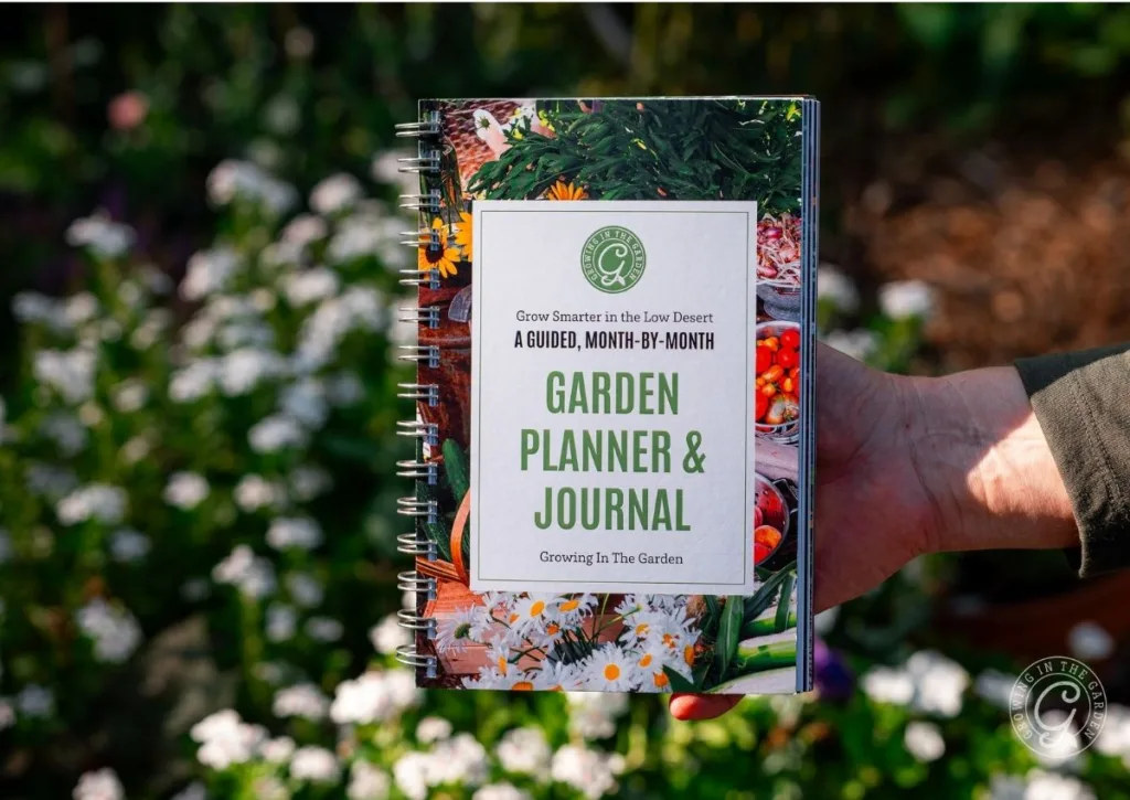 A hand holds an Arizona Garden Planner & Journal book with a floral and vegetable cover in a garden.