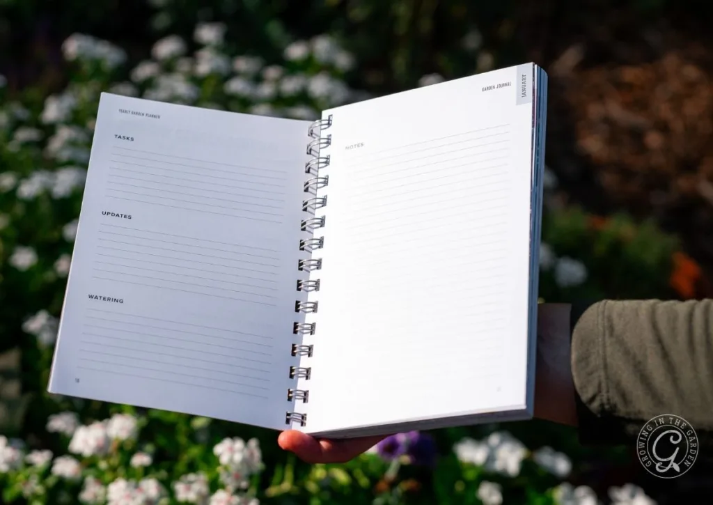 A person holds an open spiral notebook with lined pages outdoors, surrounded by blurred flowers—perfect for sketching ideas in an Arizona Garden Planner.