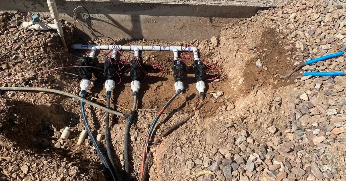 Six irrigation valves connected to pipes and wires are installed in a partially dug area with soil and rocks, making it important to carefully choose raised bed location for optimal garden layout.