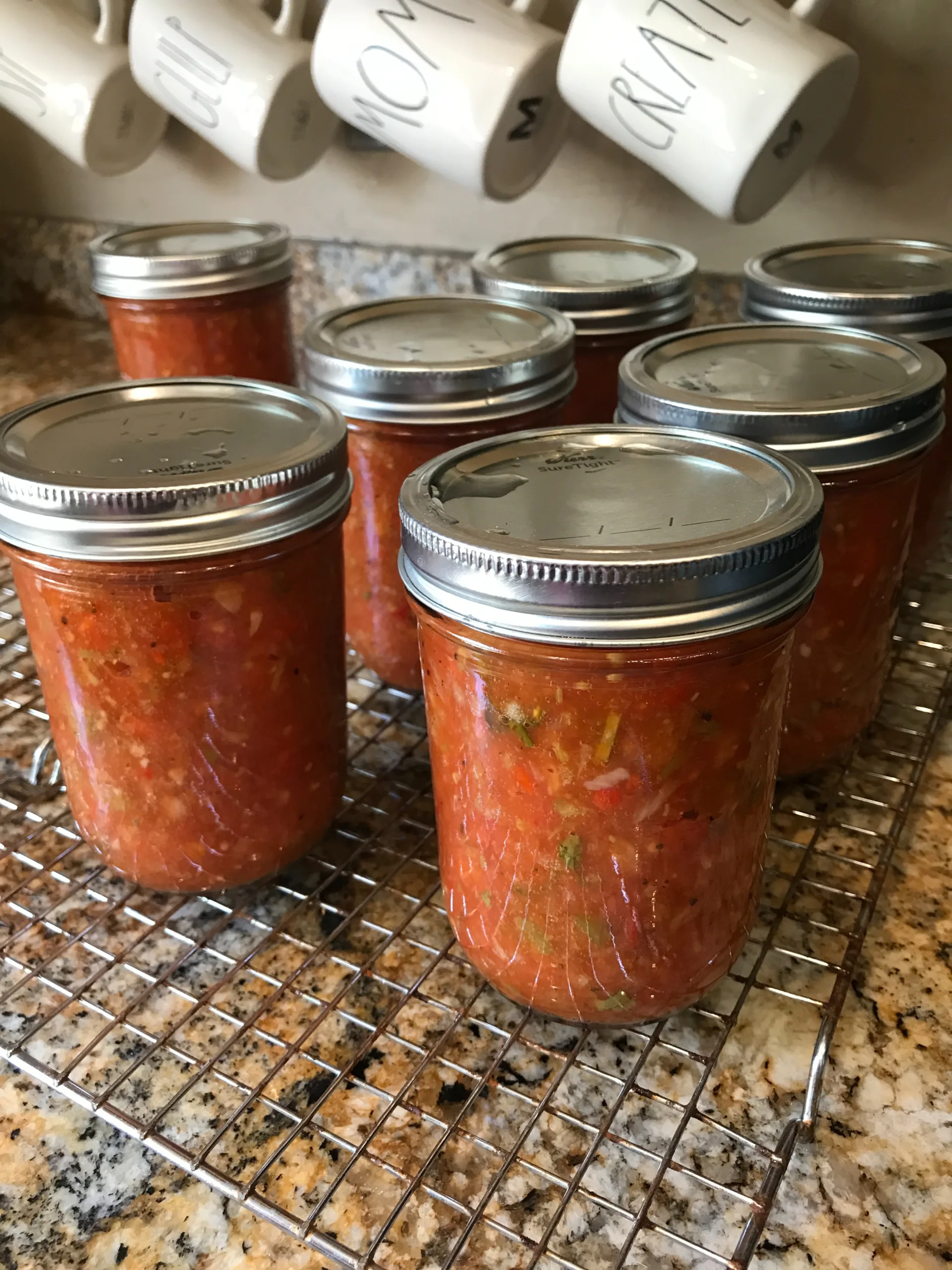 Jars of our favorite garden salsa recipe cooling on a wire rack on a granite countertop, mugs hanging above.