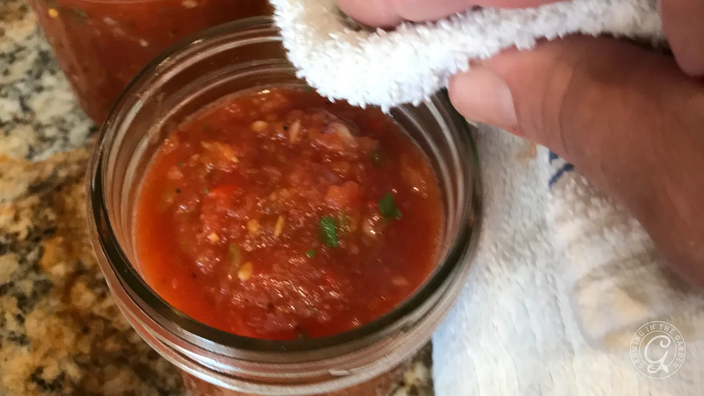 A hand wipes the rim of a glass jar filled with chunky tomato salsa using a white cloth.