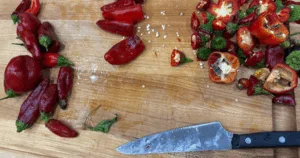 A knife and chopped roasted peppers are on a wooden cutting board, with pepper seeds scattered around.