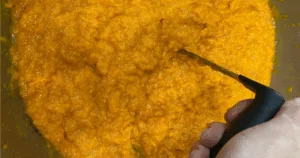 A hand holding a masher in orange mashed carrots inside a bowl, perfect for preparing a make ahead sweet potato casserole.