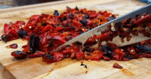 A knife slicing roasted peppers on a wooden cutting board.