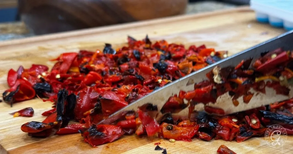 Close-up of a knife chopping roasted peppers and red chili peppers on a wooden cutting board.