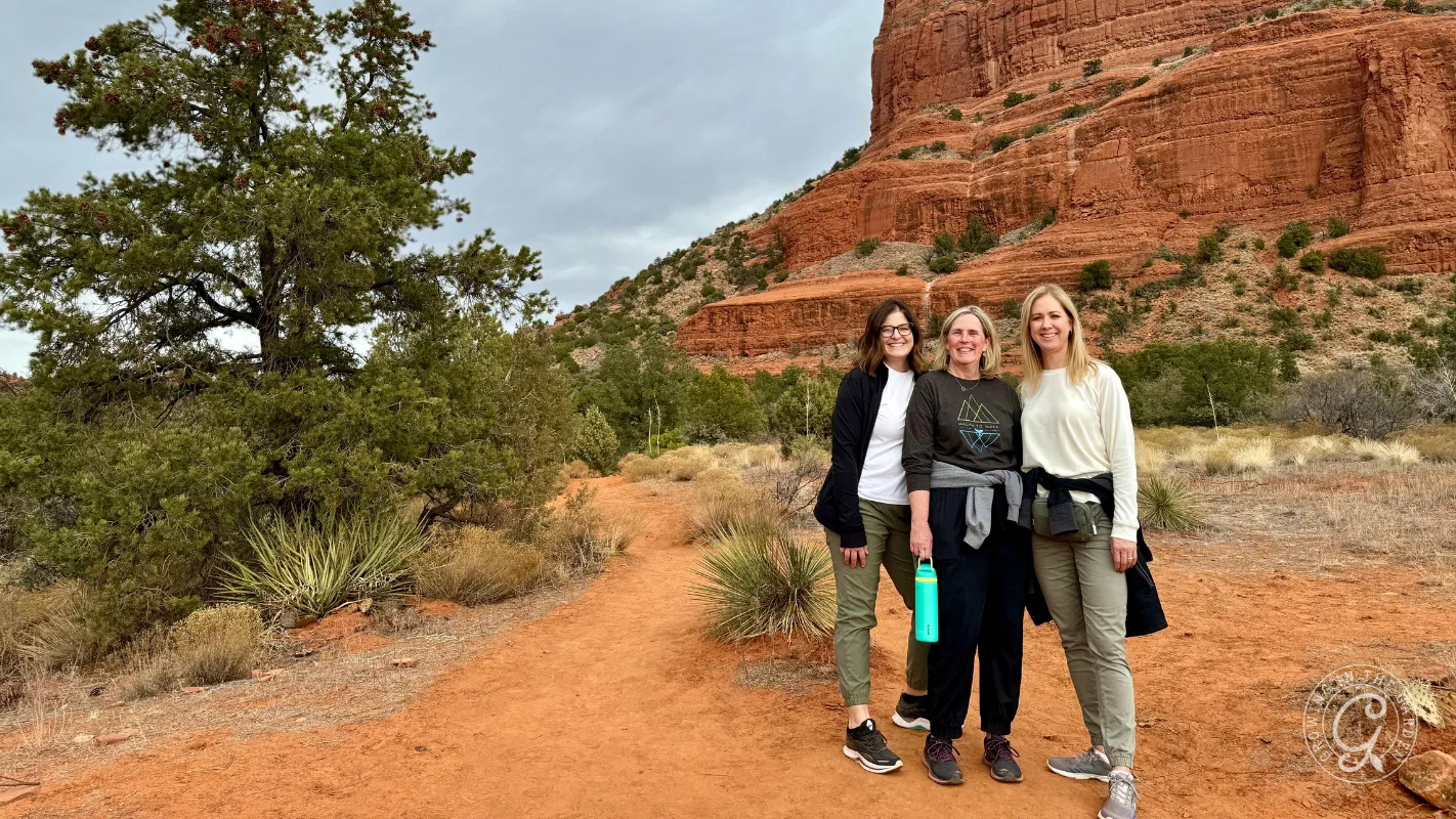 Three women smile together on a red dirt trail with red rock formations and desert plants in the background.