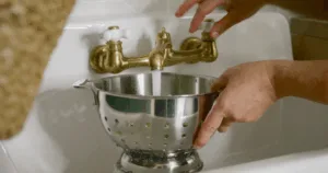 Hands holding a metal colander under a running faucet in a white sink, preparing fresh okra for an okra water recipe.