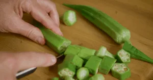 A person slicing fresh okra on a wooden cutting board with a knife, preparing to make okra water and explore its health benefits.