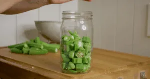 A hand places chopped okra into a glass jar on a wooden counter, preparing for making okra water&mdash;a method enjoyed by many for its potential okra water health benefits. More fresh okra and a bowl sit in the background.