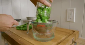 A hand pours chopped okra from a jar into a strainer over a measuring cup on a wooden counter, demonstrating how to make okra water.