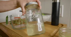 A hand holds a mason jar of cloudy okra water over a wooden counter with kitchen items in the background, highlighting the process to make okra water and enjoy its health benefits.