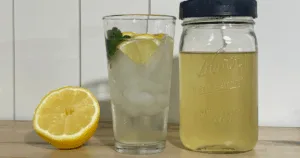 A glass of iced lemon drink, a jar of lemonade, and a halved lemon rest on a wooden surface&mdash;perfect inspiration if you're making okra water or exploring refreshing homemade beverages.
