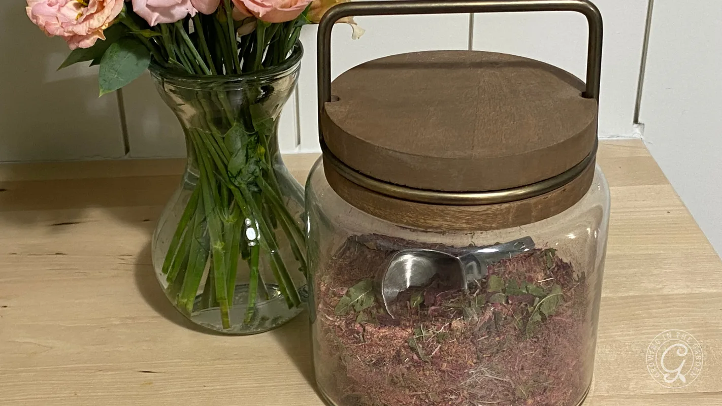Glass jar with a wooden lid and a metal scoop, next to a vase of pink flowers on a wooden surface.