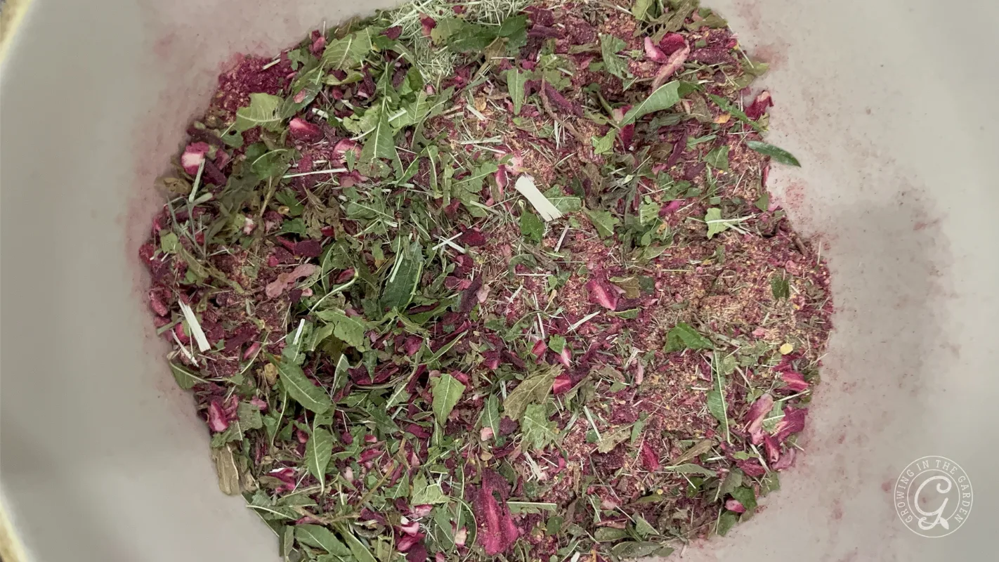 A bowl of loose, dried green and purple herbs, leaves, and petals, viewed from above.