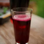 A glass of deep red juice sits on a wooden table with a blurred background.