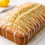 Lemon poppy seed loaf cake drizzled with glaze, with lemons in the background on a marble surface.