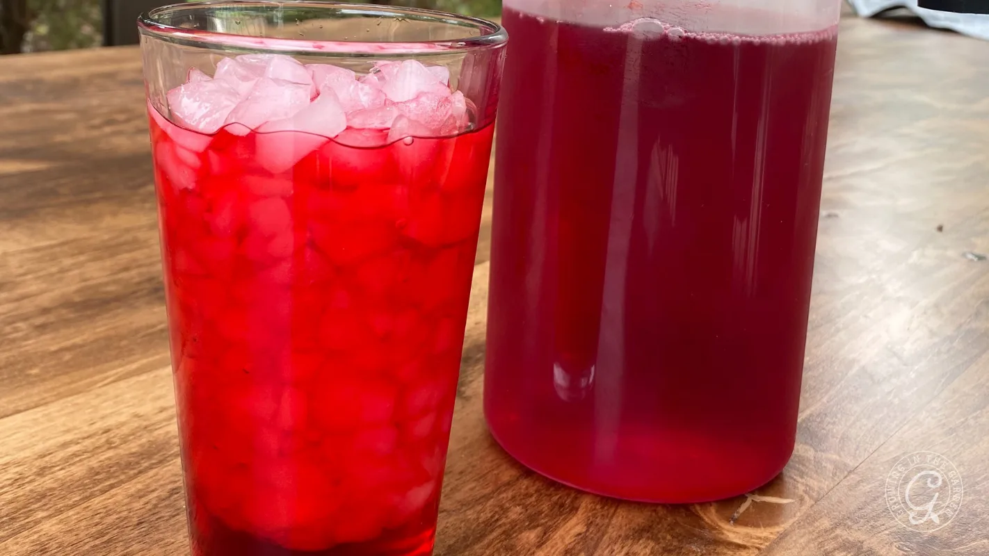 A glass of red iced drink sits next to a pitcher of the same beverage on a wooden table.