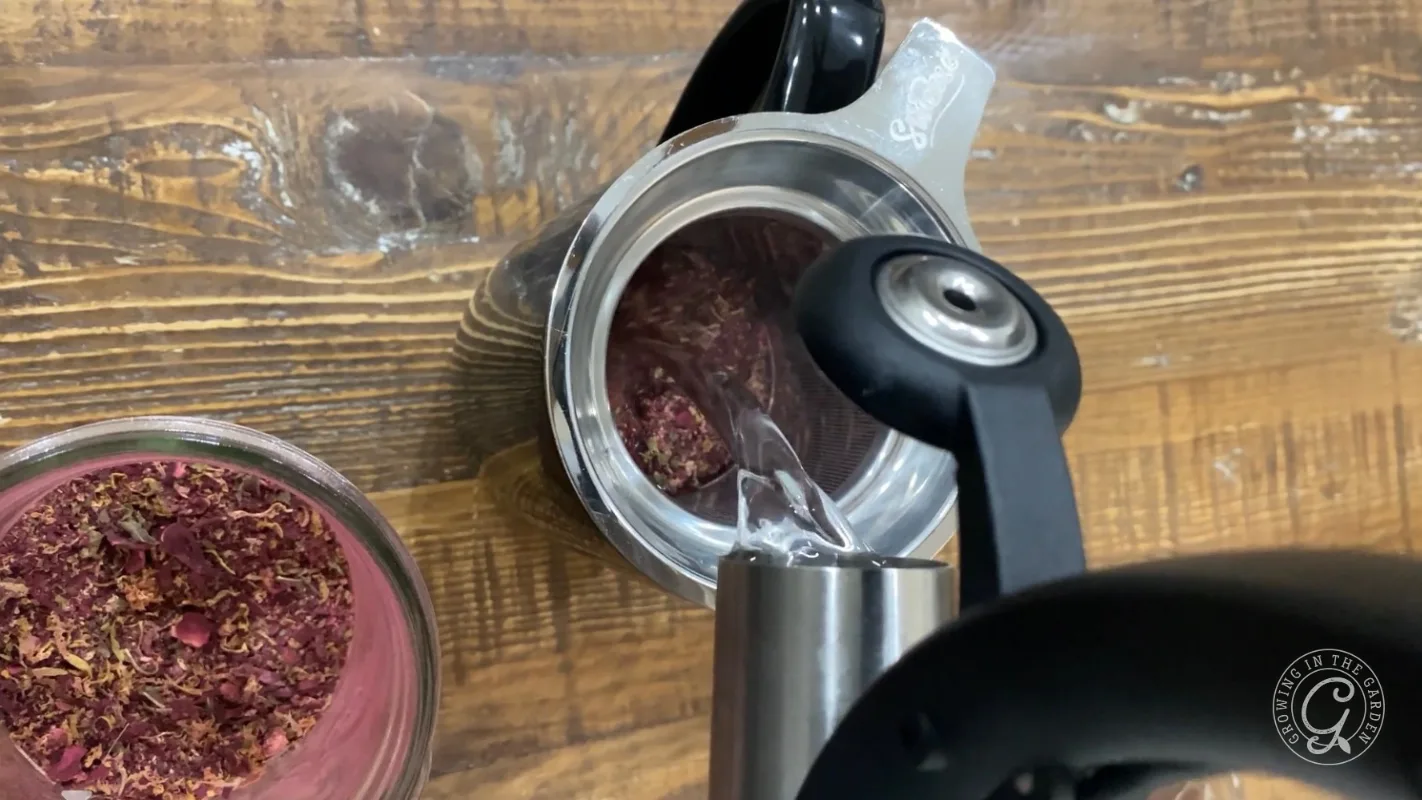 Hot water being poured over dried rose petals in a metal teapot on a wooden table, with a jar nearby.