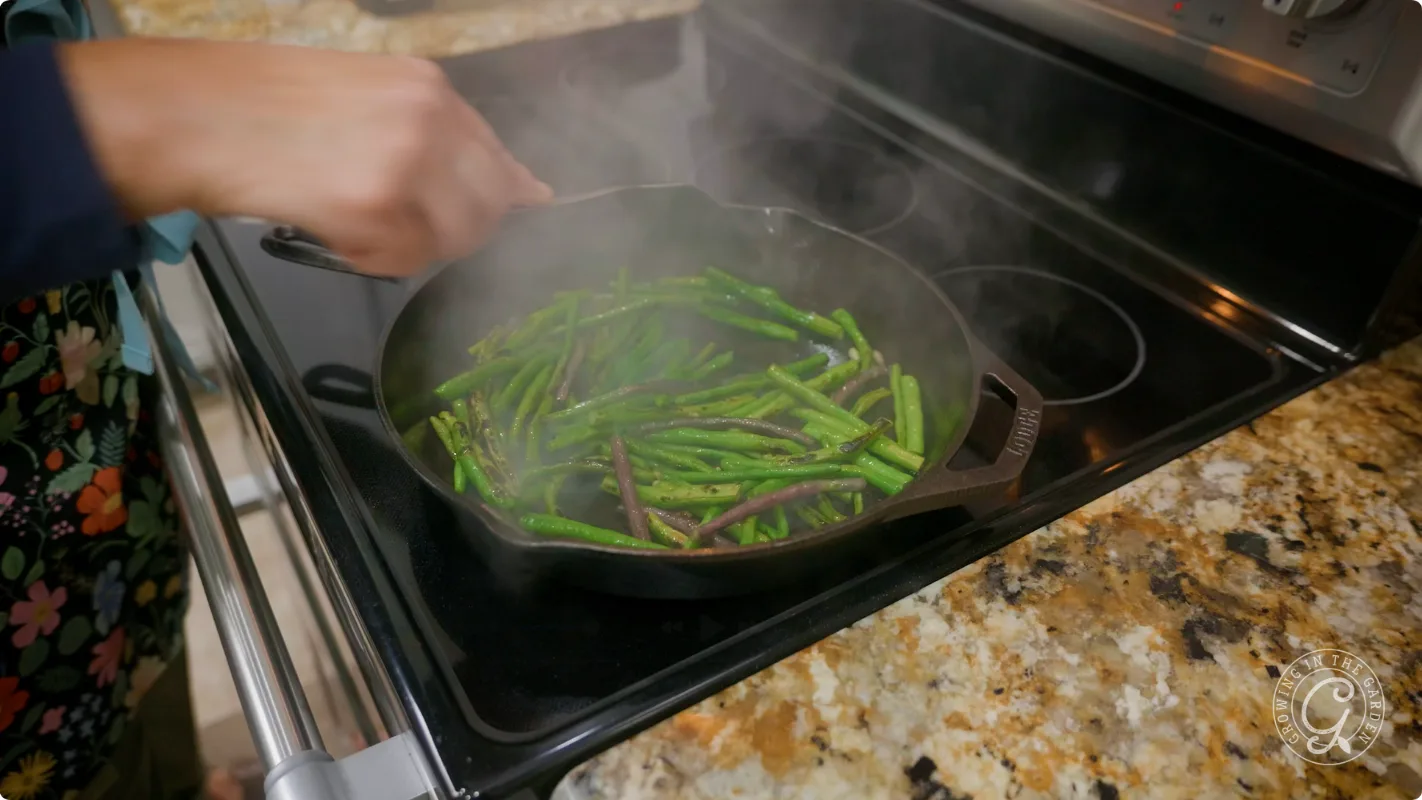A person stirs green beans in a cast iron skillet on a stovetop, with steam rising.