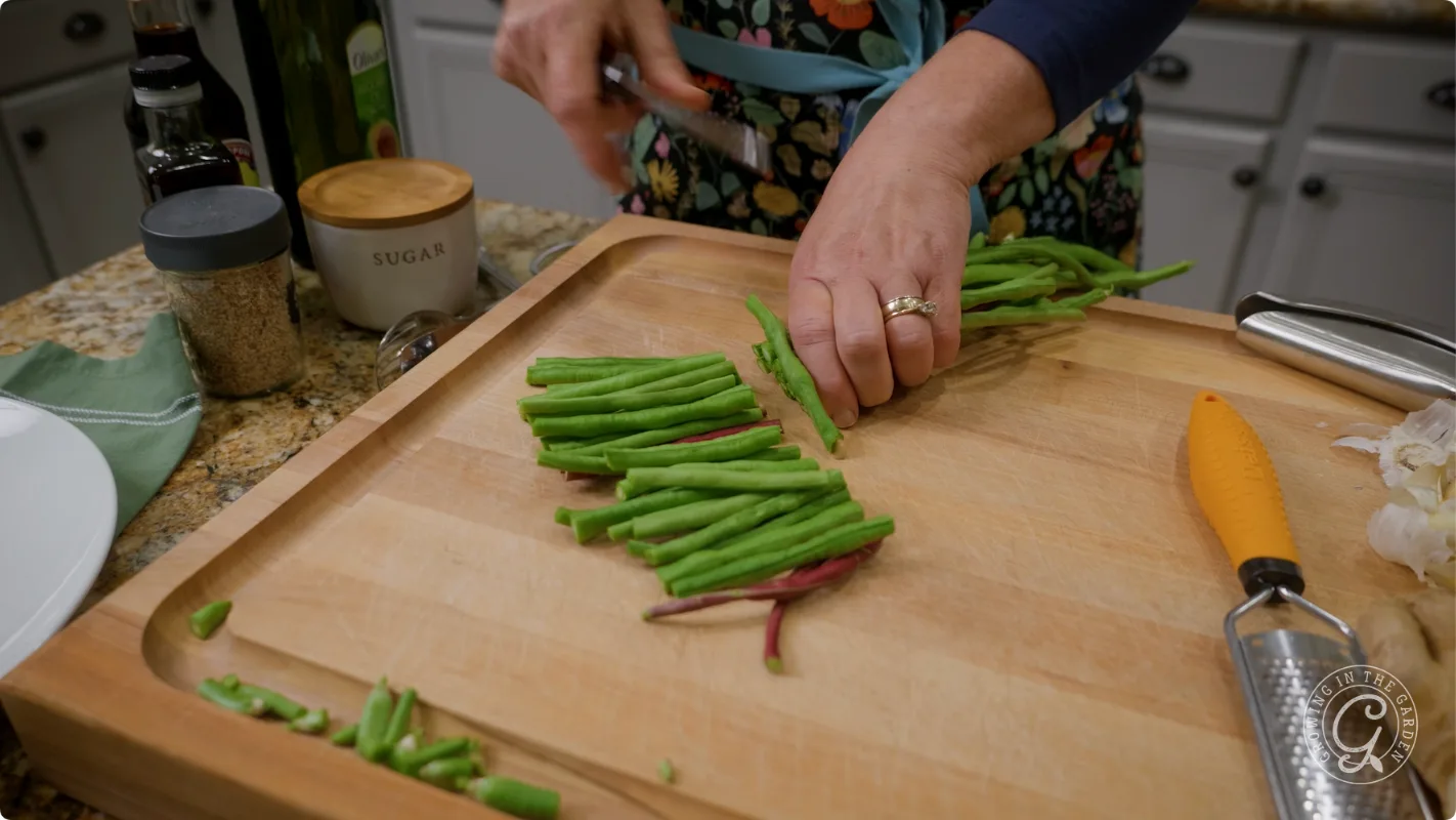 Hands chopping fresh green beans on a wooden cutting board in a kitchen with various ingredients nearby.