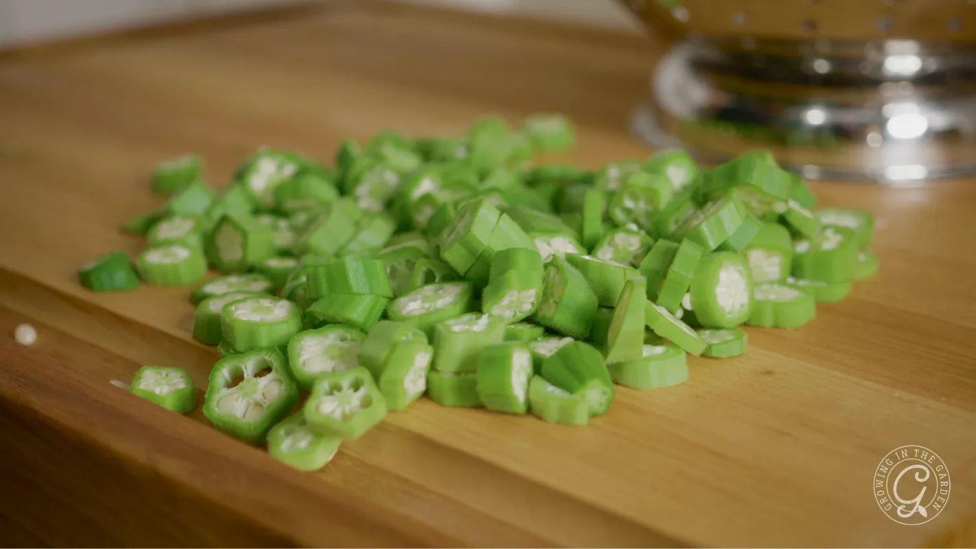Sliced okra pieces on a wooden cutting board with a metal colander in the background.
