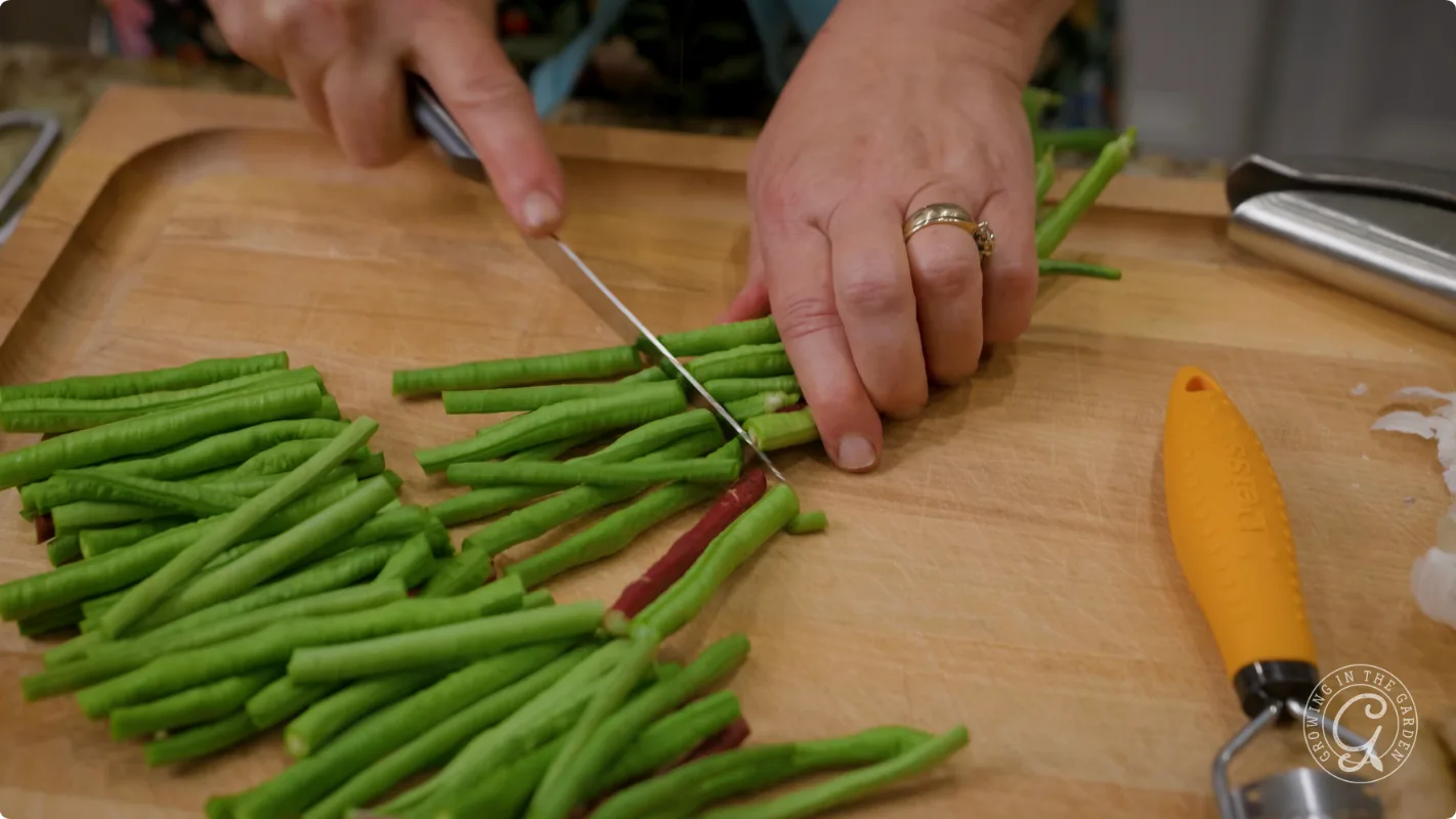 Hands slicing long green beans on a wooden cutting board with a knife; orange peeler nearby.