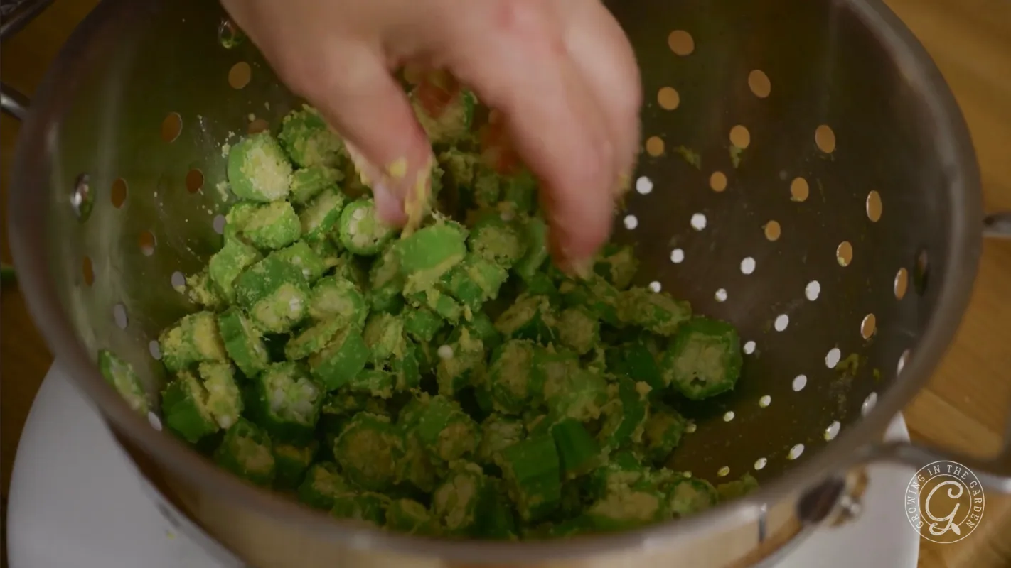A hand mixes sliced okra coated in flour or cornmeal in a metal colander.