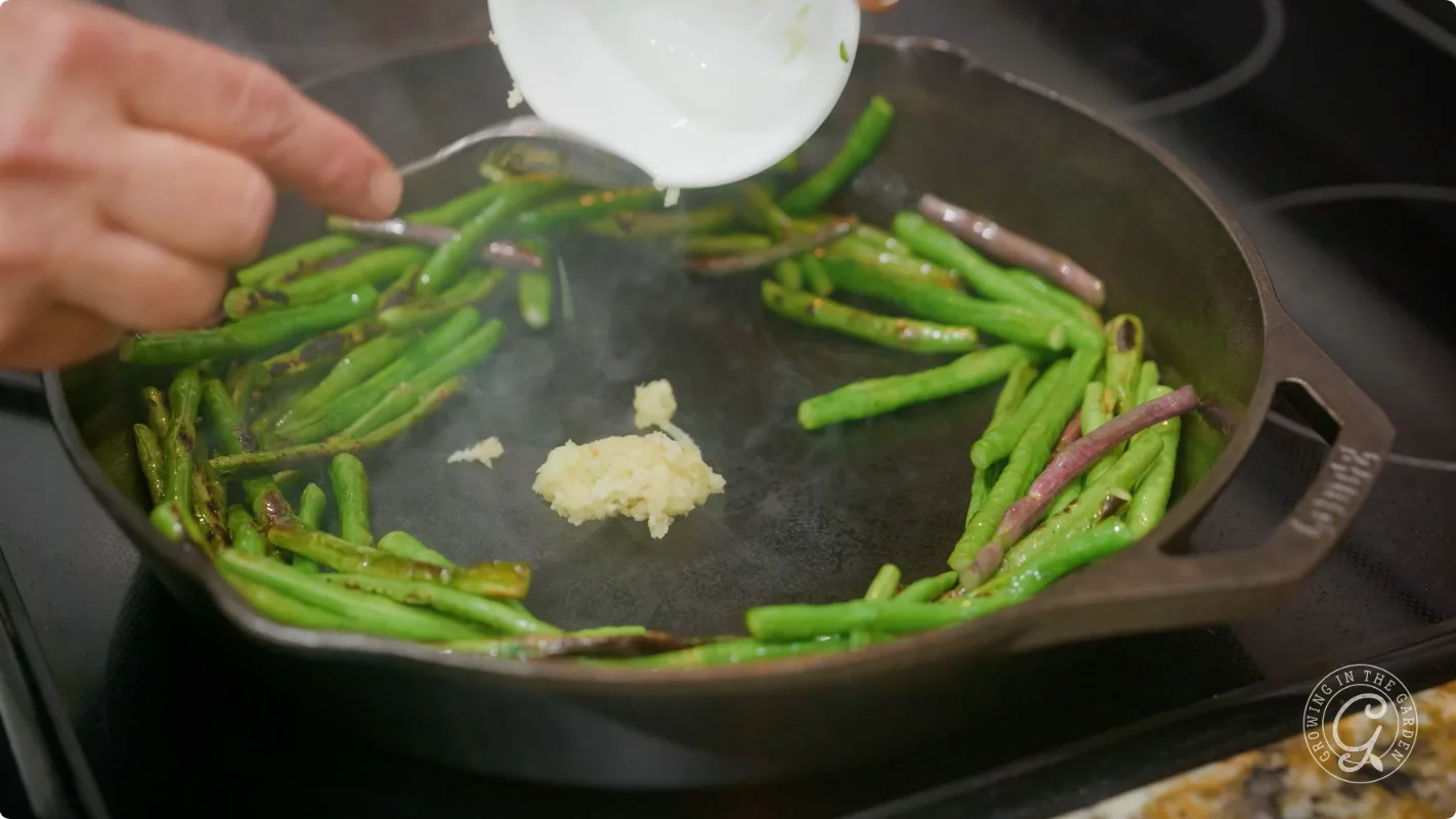 A hand adds minced garlic to a skillet with green beans cooking on a stovetop.