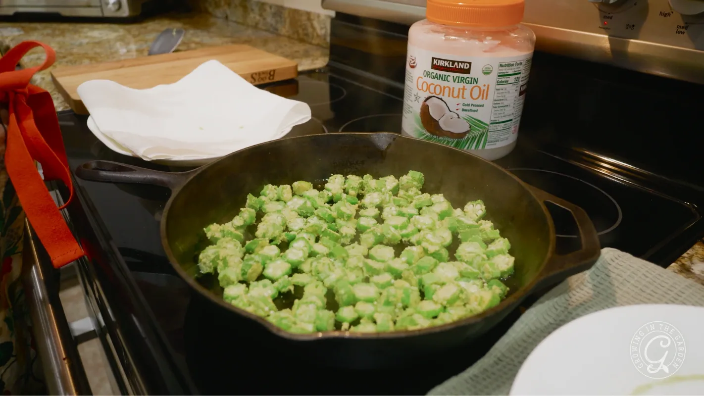 Chopped okra cooking in a cast iron skillet on a stove, with a jar of coconut oil nearby.