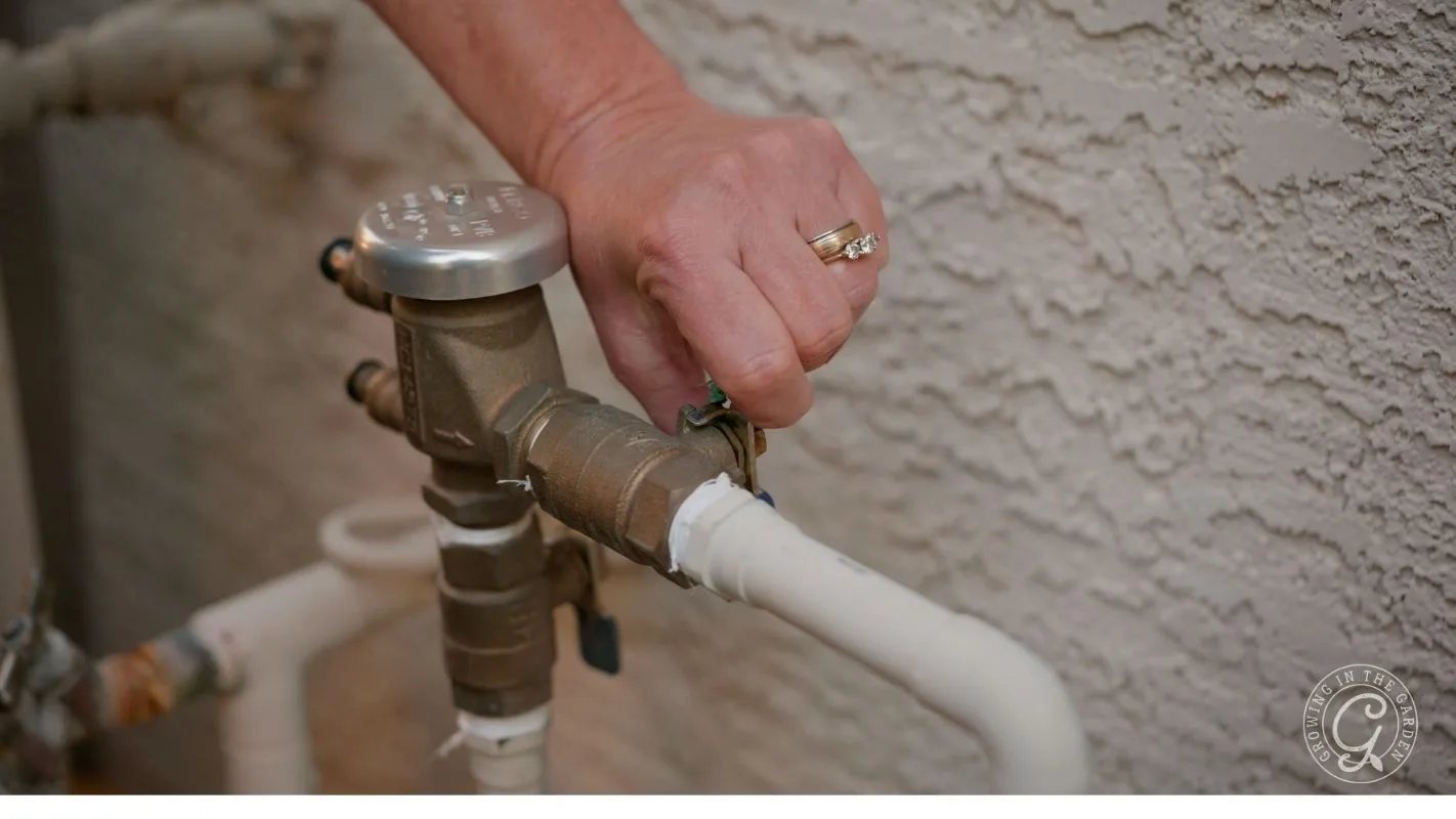 A hand turns a valve on a copper irrigation pipe, with a stucco wall in the background.