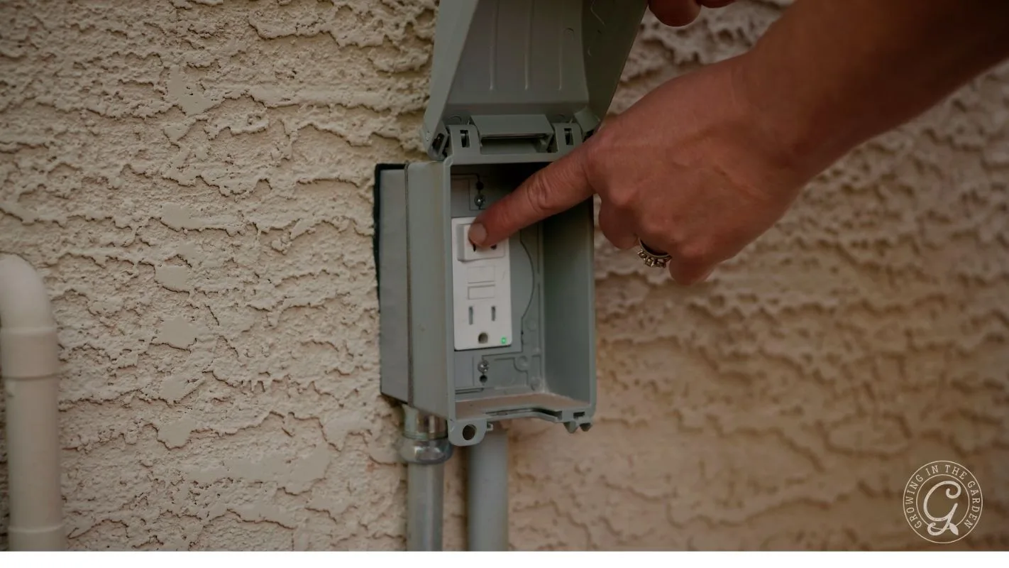 A hand pressing the test button on an outdoor electrical outlet with a protective cover.