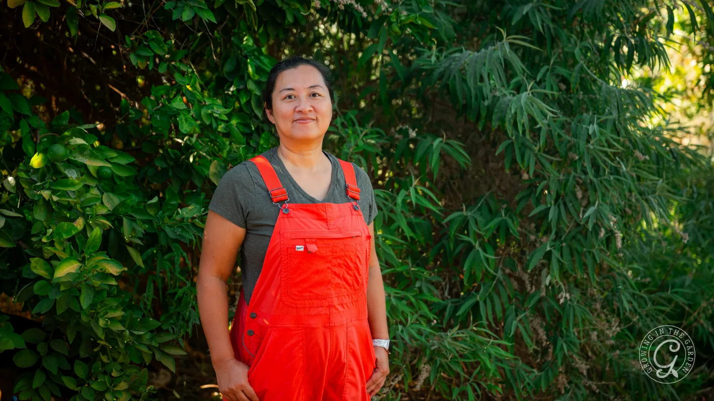 Person in a gray shirt and bright orange overalls stands outside in front of dense green foliage.